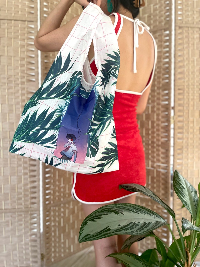 May include: A white reusable tote bag with a grid pattern and a tropical leaf print. The bag features a graphic of a person with headphones on, standing in front of a blue and purple background.