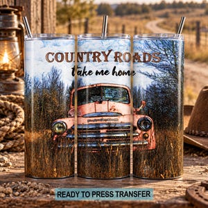 May include: A cylindrical metal tumbler featuring a graphic print of a vintage pink truck parked in a field of tall dry grass. The text "COUNTRY ROADS" and "take me home" is printed above the truck. The tumbler includes a metal straw.