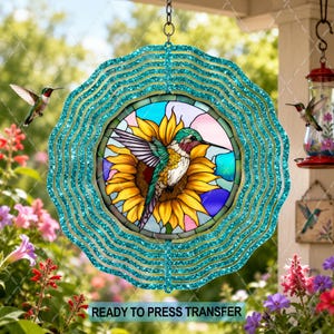 May include: A circular wind spinner featuring a glittery teal scalloped border and a central stained-glass style design. The artwork depicts a hummingbird perched on a large yellow sunflower against a backdrop of blue and pink sky segments.
