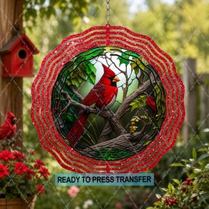 May include: A circular wind spinner featuring a vibrant red cardinal perched on a tree branch. The design is framed by a scalloped, glittery red border. The text "READY TO PRESS TRANSFER" is printed in black on a light blue rectangular background.