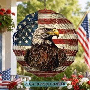 May include: A circular metal wind spinner featuring a detailed illustration of a bald eagle head with a yellow beak, set against a distressed American flag background with red, white, and blue stripes and white stars, suspended by a metal chain.