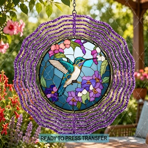 May include: A circular wind spinner featuring a purple glittery frame with a central stained glass-style insert. The insert depicts a hummingbird with blue and green feathers perched on a branch surrounded by pink and purple flowers.