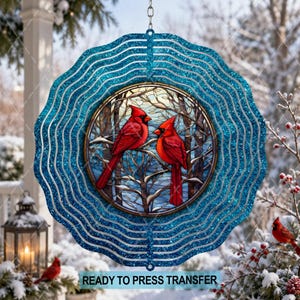 May include: A circular wind spinner featuring a glittery blue wavy border and a central stained glass-style insert. The insert depicts two bright red cardinals perched on snow-covered tree branches against a pale, wintry sky background.