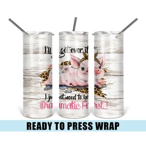 May include: A white tumbler with a woodgrain background features a pink pig with a leopard print blanket. The text on the tumbler reads "I'll get over it I just need to be dramatic first!"