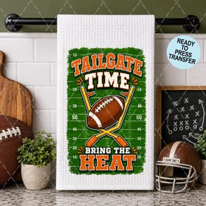 May include: A white waffle-weave kitchen towel featuring a green football field graphic with yard lines, a brown football, yellow goalposts, and the orange and white text "TAILGATE TIME BRING THE HEAT" printed in the center of the design.