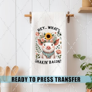 May include: White kitchen towel with a cartoon pig wearing a floral crown and a plaid scarf. The text reads "HEY... WHAT'S SHAKIN' BACON?" and "READY TO PRESS TRANSFER". The towel is hanging on a wooden bar.