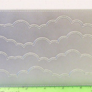 Pode incluir: Um die-cut de metal prateado com um design de quatro filas de nuvens festoneadas. O die-cut mede aproximadamente 12,7 cm de largura e 10,2 cm de altura.