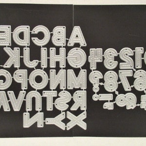 May include: A collection of silver metal alphabet and number die cuts. The letters and numbers are in a bold, sans-serif font with a dotted line detail. The die cuts are arranged on a black background.