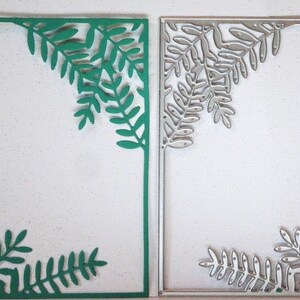 Op de afbeelding: Twee rechthoekige papieren uitsparingen met bladeren. Eén is groen, de andere zilver. De bladeren bevinden zich in de hoeken van de rechthoeken. De achtergrond is wit met kleine spikkels.