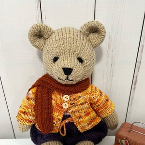 May include: A knitted teddy bear wearing a brown scarf, orange and yellow sweater, purple pants, and orange knitted shoes. The bear is sitting next to a small suitcase and a knitted orange pumpkin.