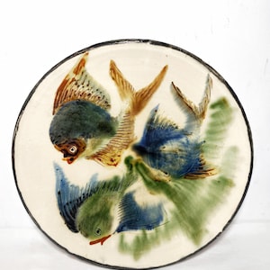 May include: Round ceramic plate with a cream-colored background and a black rim. The plate features a hand-painted design of three colorful fish in shades of blue, green, and brown, with green aquatic plants.