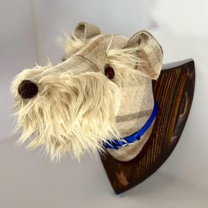 Puede incluir: Cabeza de perro decorativa hecha de tela beige con una barba desgreñada de color crema y ojos de botón marrones. La cabeza del perro está montada en una placa de madera marrón oscuro y lleva un collar azul.