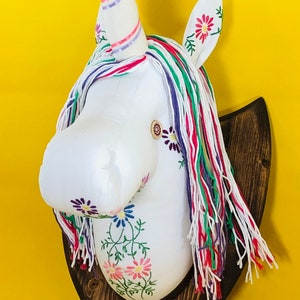May include: White unicorn head with a pink, purple and green yarn mane and tail. The unicorn has a pink and white striped horn and is embroidered with flowers. The head is mounted on a dark brown wooden plaque.