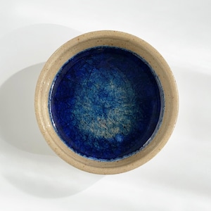 Könnte beinhalten: Eine runde Keramikschale mit strukturiertem, tiefblauem Innenraum und beigem Rand. Die Schale befindet sich auf einer weißen Oberfläche, wobei Schatten Tiefe verleihen. Das Design ist schlicht und elegant.