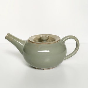 May include: A sage green ceramic teapot with a matching lid. The teapot has a rounded body, a curved spout, and a loop handle. The lid is a lighter shade of green with a small knob. The teapot is on a white background.