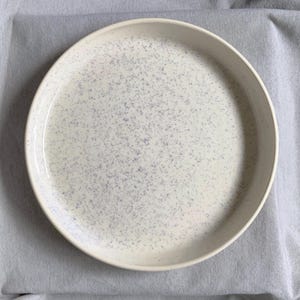 May include: A round ceramic plate with a smooth, off-white base colour and a speckled pattern of small, light purple dots distributed across the entire surface. The plate features a slightly raised rim and a clean, circular design.