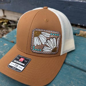 Könnte beinhalten: Braune Trucker-Cap mit weißem Mesh-Rücken und gebogenem Schirm. Auf der Vorderseite befindet sich ein Lederpatch mit einem Gänseblümchen-Design in Weiß und Gold, akzentuiert mit türkisfarbenen Punkten. Die Kappe hat ein Richardson 115 Trucker-Etikett.