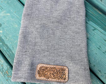 May include: Gray beanie with a brown leather patch featuring a carved floral design.