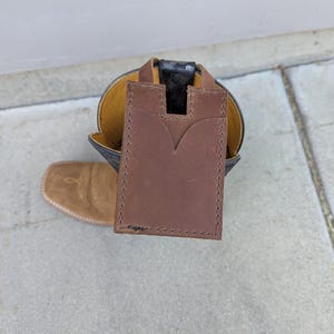 Könnte beinhalten: Ein brauner Lederkartenhalter mit einem V-förmigen Ausschnitt liegt vor einem schwarzen und gelben Cowboy-Stiefel. Der Kartenhalter ist rechteckig mit genähten Kanten. Der Stiefel hat eine spitze Spitze.