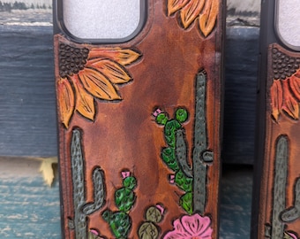 May include: A brown leather phone case with a carved design of sunflowers, cacti, and pink flowers. The case has a cutout for the phone's camera.