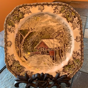 May include: Square ceramic plate with a winter landscape design. The plate depicts a covered bridge, trees, and a snowy scene in brown, green, and white. The rim has a decorative floral and leaf pattern. The plate is displayed on a black metal stand.