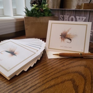 May include: A stack of cream-colored greeting cards with a gold border and a watercolor illustration of a fishing fly. One card is displayed upright, showing the fly and the text "Black Adams Caddis". A wooden pen rests on the table.