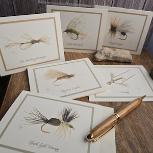 May include: A collection of greeting cards featuring watercolor illustrations of various fishing flies. Each card has a cream-colored background with a gold border and the fly's name printed below. A wooden pen with gold accents rests on the wooden surface.
