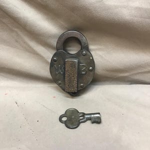 May include: A vintage heart-shaped padlock with a key. The lock is made of tarnished metal and features the initials "W B". The key is placed next to the lock. The lock has a textured, cork-like material in the center.