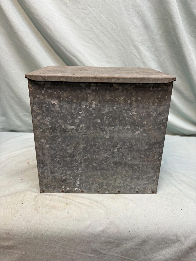 Peut inclure: Un bac de rangement en m&eacute;tal galvanis&eacute; avec un couvercle &agrave; charni&egrave;re. Le bac a une forme rectangulaire et une finition gris argent&eacute; mouchet&eacute;. Le couvercle est d'une nuance de gris plus claire. L'objet semble vintage ou antique.