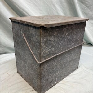Peut inclure: Un conteneur de stockage en m&eacute;tal galvanis&eacute; avec un couvercle et une poign&eacute;e en fil de fer. La bo&icirc;te rectangulaire a un aspect l&eacute;g&egrave;rement vieilli, avec des coutures et des rivets visibles. Le couvercle est bien ajust&eacute; et la poign&eacute;e est arqu&eacute;e.