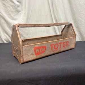 May include: A vintage metal tool tote with a weathered finish and a central carrying handle. The side features the red printed text WEN TOTER inside a red rectangular graphic. The metal shows signs of oxidation and wear across its surface.