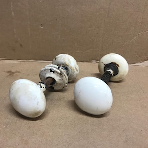 May include: A collection of five antique white porcelain door knobs. Each knob has a rounded, egg-like shape with a dark metal stem. Some knobs show signs of age and wear, with visible discoloration and chipping.