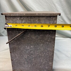 Peut inclure: Un r&eacute;cipient rectangulaire en m&eacute;tal galvanis&eacute; avec un couvercle. Le r&eacute;cipient mesure environ 23 cm de large. Il a une poign&eacute;e et est rivet&eacute;. Le m&eacute;tal a un aspect mouchet&eacute;.