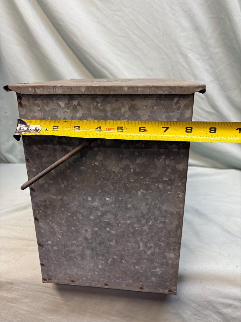 Peut inclure: Un r&eacute;cipient rectangulaire en m&eacute;tal galvanis&eacute; avec un couvercle. Le r&eacute;cipient mesure environ 23 cm de large. Il a une poign&eacute;e et est rivet&eacute;. Le m&eacute;tal a un aspect mouchet&eacute;.