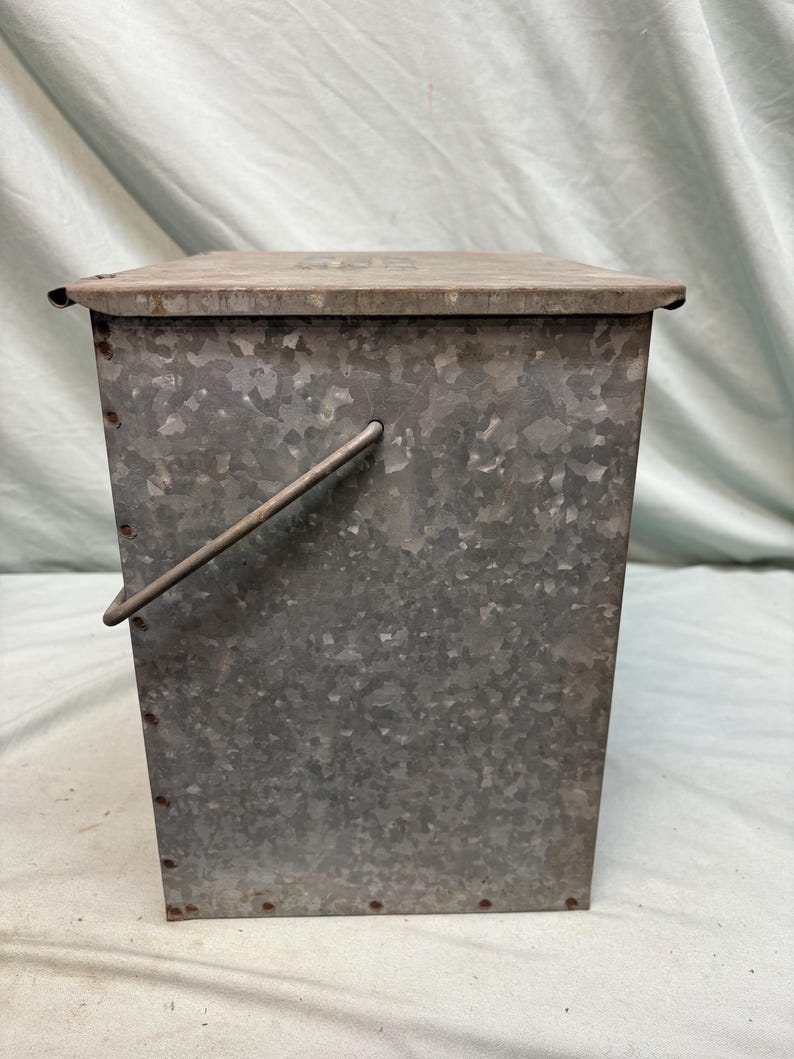 Peut inclure: Un conteneur en m&eacute;tal galvanis&eacute; avec un couvercle et une poign&eacute;e. Le conteneur rectangulaire a une surface textur&eacute;e et est rivet&eacute;. Le couvercle est pos&eacute; &agrave; plat sur le dessus. La poign&eacute;e est en m&eacute;tal et est fix&eacute;e sur le c&ocirc;t&eacute;.