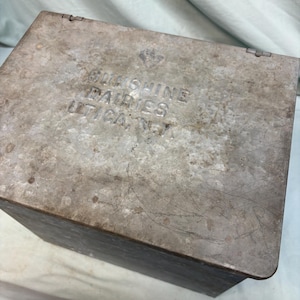 Peut inclure: Une bo&icirc;te m&eacute;tallique rectangulaire vintage avec un couvercle &agrave; charni&egrave;re. La bo&icirc;te pr&eacute;sente une finition grise patin&eacute;e avec des rayures et une usure visibles. Le texte "Sunshine Dairies Utica, N.Y." est estamp&eacute; sur le couvercle.