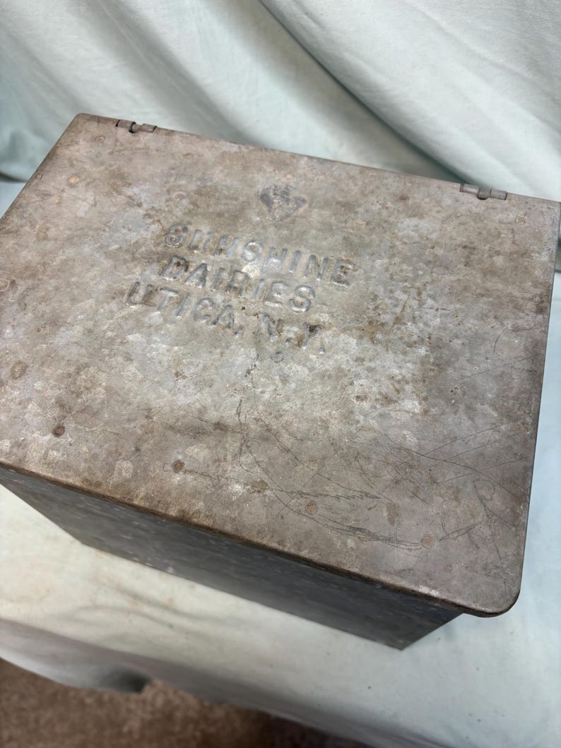 Peut inclure: Une bo&icirc;te m&eacute;tallique rectangulaire vintage avec un couvercle &agrave; charni&egrave;re. La bo&icirc;te pr&eacute;sente une finition grise patin&eacute;e avec des rayures et une usure visibles. Le texte "Sunshine Dairies Utica, N.Y." est estamp&eacute; sur le couvercle.