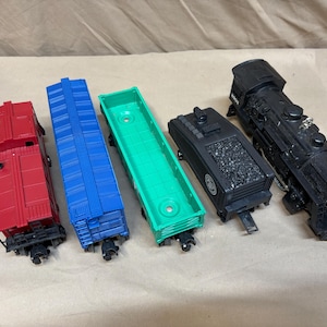 May include: A collection of toy train cars in various colors. The set includes a red caboose, a blue boxcar, a green flatcar, a black coal car, and a black steam engine. The toy train cars are arranged on a neutral surface.