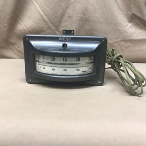 May include: A gray vintage West temperature gauge with a curved face and a range from 5 to 25. The gauge has a green cord attached and the word "WEST" is printed on the top.