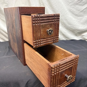 May include: A dark brown wooden storage unit featuring two stacked drawers. Each drawer front displays a carved grid pattern and a small, round, metallic floral-shaped knob. The wood grain is visible, and the drawers are pulled partially open.
