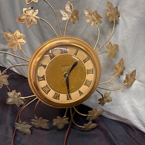 May include: A gold-toned wall clock featuring a circular face with Roman numerals and the word United printed in the center. The clock is surrounded by radiating metal arms, each tipped with a textured gold-colored leaf-shaped ornament.