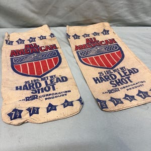 May include: Two rectangular off-white canvas bags featuring the text ALL AMERICAN, 25 LBS. NET WT. HARD LEAD SHOT, and RSR CORPORATION PRODUCT. The bags display a red, white, and blue shield graphic with stars and the number 7 1/2 printed on them.