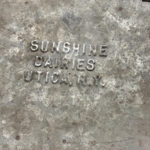 Peut inclure: Gros plan sur un r&eacute;cipient m&eacute;tallique vieilli avec le texte en relief "SUNSHINE DAIRIES UTICA, N.Y.". Le r&eacute;cipient pr&eacute;sente un aspect us&eacute; et vieilli avec des rayures et imperfections visibles, sugg&eacute;rant une utilisation vintage.