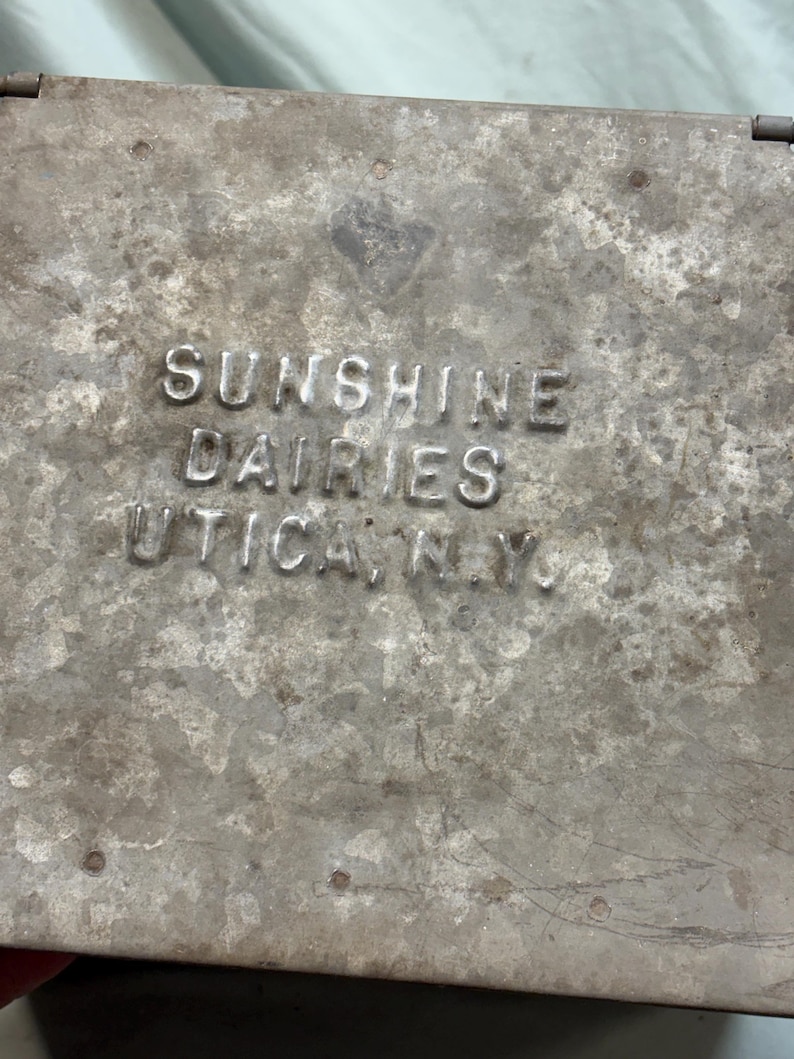 Peut inclure: Gros plan sur un r&eacute;cipient m&eacute;tallique vieilli avec le texte en relief "SUNSHINE DAIRIES UTICA, N.Y.". Le r&eacute;cipient pr&eacute;sente un aspect us&eacute; et vieilli avec des rayures et imperfections visibles, sugg&eacute;rant une utilisation vintage.