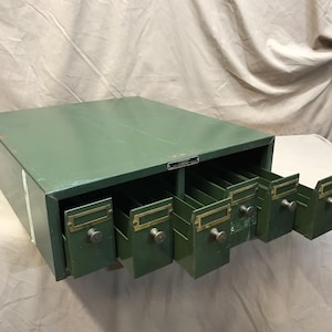 May include: Vintage green metal file cabinet with multiple drawers. Each drawer has a brass label holder and a round metal knob. The cabinet is a muted green color and shows signs of wear.