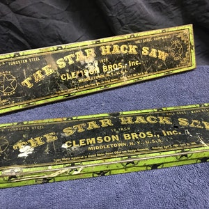 May include: Two vintage metal boxes for "The Star Hack Saw" hand saws. The boxes are black with gold lettering and a green border. The text includes "Tungsten Steel" and "Clemson Bros., Inc." The saws are 10 inches long.