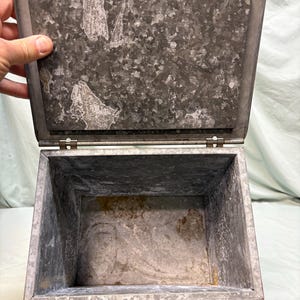 Peut inclure: Une bo&icirc;te m&eacute;tallique rectangulaire ouverte avec un couvercle &agrave; charni&egrave;re. La bo&icirc;te est en acier galvanis&eacute; avec un aspect mouchet&eacute; et vieilli. Le couvercle est l&eacute;g&egrave;rement entrouvert, r&eacute;v&eacute;lant l'int&eacute;rieur vide. Une main maintient la bo&icirc;te ouverte.