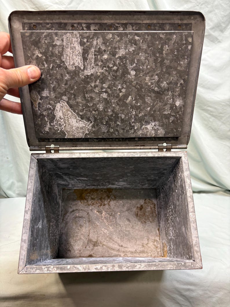 Peut inclure: Une bo&icirc;te m&eacute;tallique rectangulaire ouverte avec un couvercle &agrave; charni&egrave;re. La bo&icirc;te est en acier galvanis&eacute; avec un aspect mouchet&eacute; et vieilli. Le couvercle est l&eacute;g&egrave;rement entrouvert, r&eacute;v&eacute;lant l'int&eacute;rieur vide. Une main maintient la bo&icirc;te ouverte.