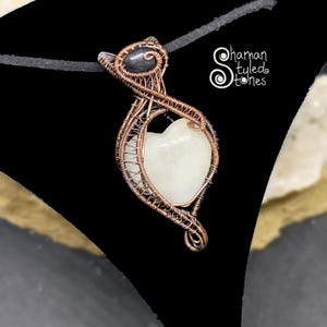 Peut inclure: Pendentif artisanal avec une pierre claire en forme de cœur, sertie dans du fil de cuivre. Le pendentif a une pierre noire en haut et est suspendu à un cordon noir. Le texte "Shaman Styled Stones" est visible à droite.