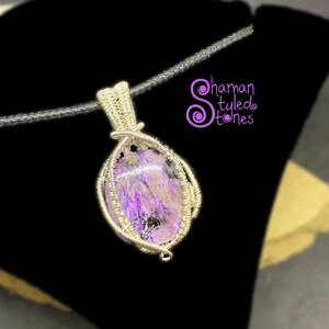 May include: A silver wire wrapped pendant featuring a purple and white stone. The pendant is hanging from a black cord.